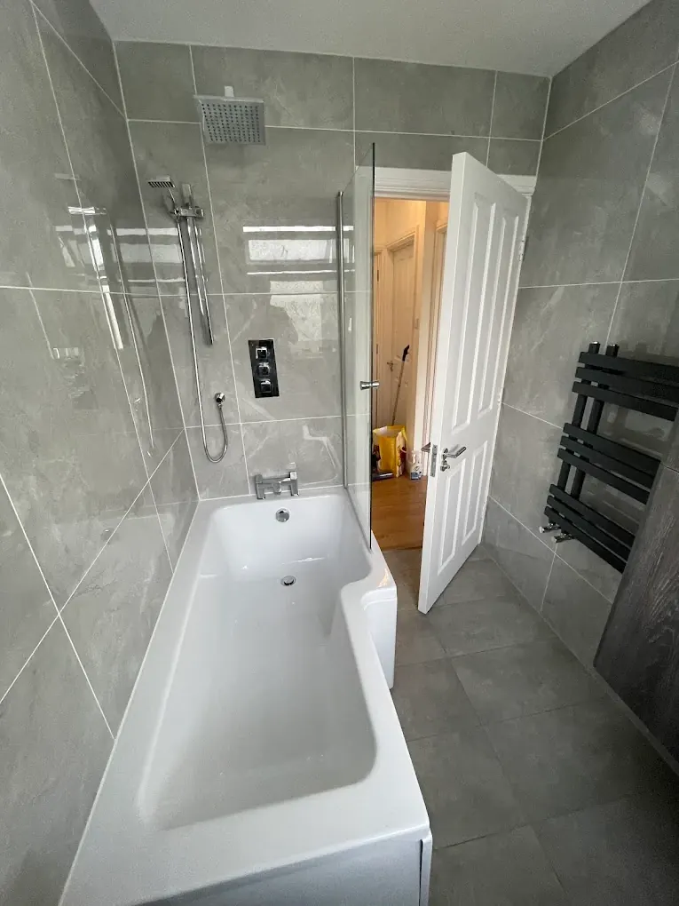 Bath with shower over and glass screen in large-format grey tiles, black towel radiator behind